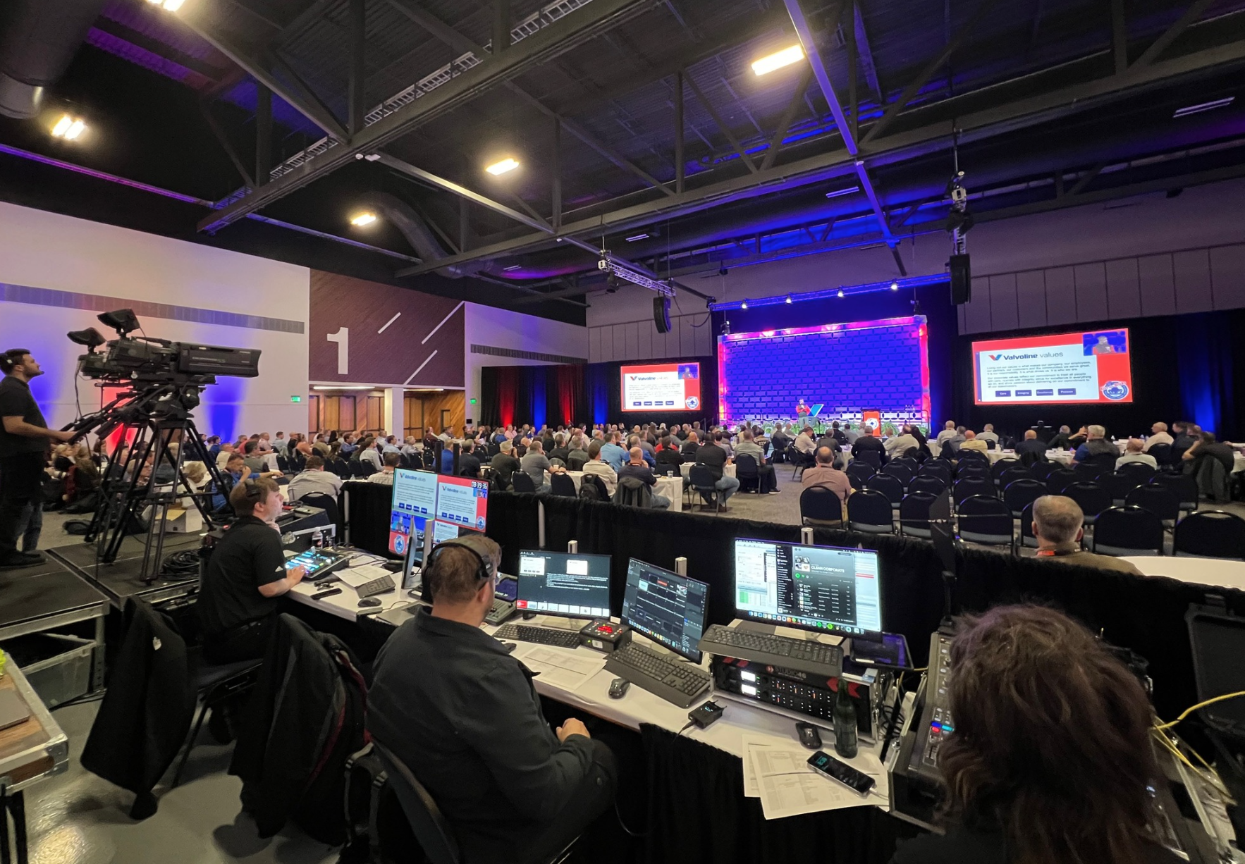 Videography, sound, and tech area/set up in the back of a large banquet room, with tech crews running tech during a corporate conference.